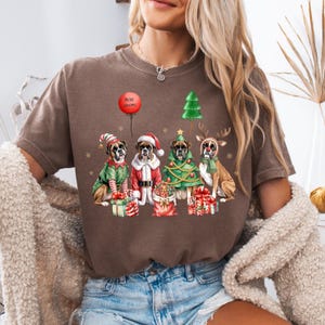 Pu&ograve; includere: T-shirt marrone con grafica natalizia. Raffigura quattro cani Boxer vestiti con abiti festivi: un elfo, Babbo Natale, un albero di Natale e una renna. Un palloncino rosso mostra le parole "Merry Christmas". Sono presenti anche regali e un albero di Natale.
