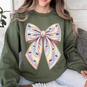 May include: An olive green sweatshirt featuring a large, iridescent bow design. The bow is adorned with small, cartoon ghost figures. The bow has a gold glitter outline. The sweatshirt is a casual, festive garment.