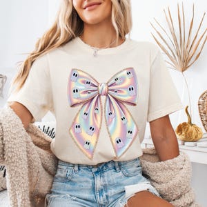 May include: Cream-colored t-shirt featuring a large, iridescent bow design adorned with cartoon ghost figures. The bow has a gradient of pastel colors, including pink, yellow, and blue. The ghosts have black eyes and mouths. The shirt is a casual, everyday wear item.