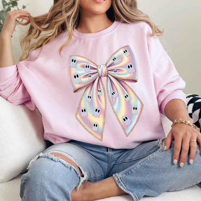May include: A light pink sweatshirt featuring a large, iridescent bow with ghost illustrations. The bow has a gold outline and is the focal point of the design. The sweatshirt is a long-sleeved crew neck.