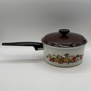 Puede incluir: Una cacerola vintage con tapa marrón y asa negra. La olla blanquecina presenta un colorido diseño de frutas y verduras alrededor de la base. La tapa tiene un pomo negro. Un artículo de menaje de cocina clásico.