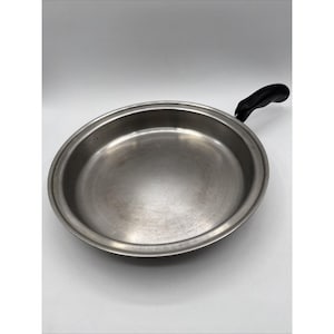 Puede incluir: Una sartén de acero inoxidable con un mango negro. La sartén es redonda con un borde ligeramente inclinado. El mango es curvo y está unido al lateral de la sartén. La sartén está diseñada para cocinar.