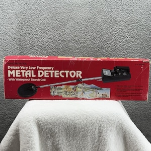 May include: A vintage Micronta Deluxe Very Low Frequency Metal Detector in its original box. The red box features the product name and a graphic of people using the detector. The detector has a black search coil and control panel.