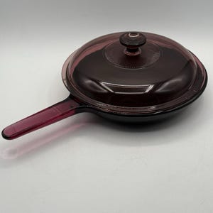Puede incluir: Una sartén de vidrio morado oscuro con una tapa a juego. La sartén tiene un mango largo y una superficie de cocción redonda. La tapa tiene un diseño circular y un pequeño pomo. El utensilio de cocina es de vidrio.