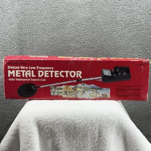 May include: A red box with the words "MICRONTA METAL DETECTOR" printed on it. The box features a diagram of the metal detector and its components. The metal detector has a black search coil and a silver handle.
