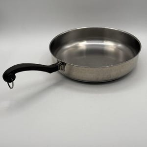 May include: Stainless steel frying pan with a black handle and a small metal loop at the end. The pan has a wide, shallow cooking surface and a slightly curved rim. The handle is ergonomically designed for a comfortable grip.