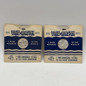 May include: Two vintage View-Master reel packages. Each package is cream-colored with blue and white text and graphics. The text reads "ONE VIEW-MASTER REEL" and "7 MORE WONDERS OF THE WORLD". One reel is labeled "344 Howe Caverns New York I". The other is labeled "SP-9022 Howe Caverns - II".