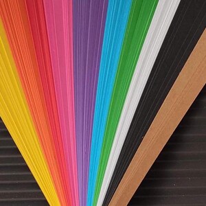 10 Colors Quilling Paper Strips - High Quality 180 gsm Rainbow Palette Set DIY Kit, Typography & 3D Effect, Perfect Craft Supplies. Quilling paper strips in various widths, including 3mm, 5mm, 7mm, and 10mm. The paper is 180 GSM and comes in a pack of 100 strips. Best for - On-edge quilling, Lettering, Outline quilling and Structured designs'.The colors are black, brown, white, green, blue, purple, pink, red, orange, and yellow. The strips are arranged in a vertical row.