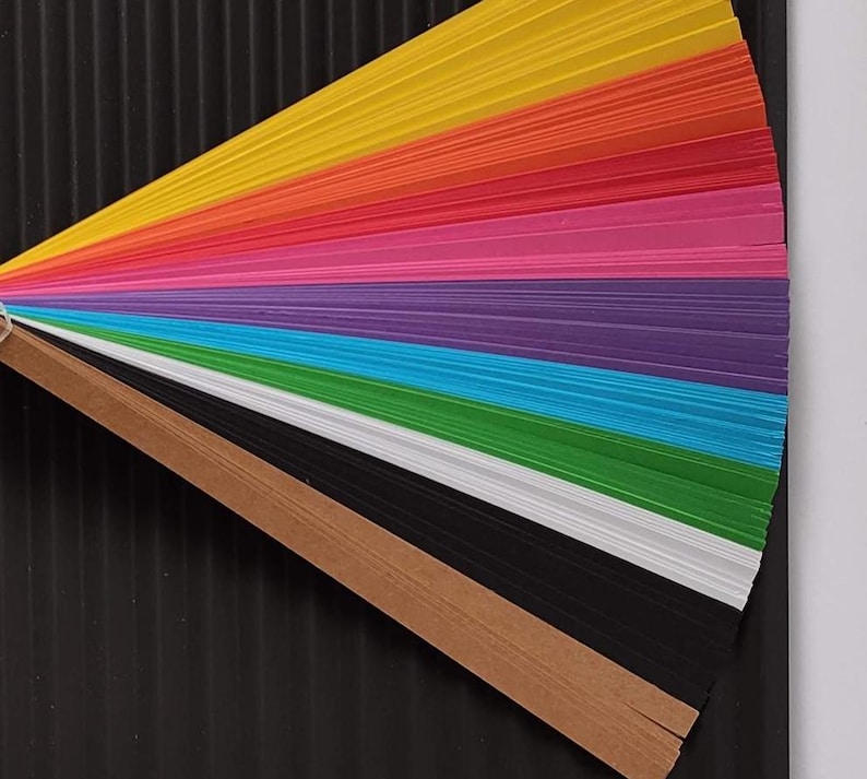 10 Colors Quilling Paper Strips - High Quality 180 gsm Rainbow Palette Set DIY Kit, Typography & 3D Effect, Perfect Craft Supplies. Quilling paper strips in various widths, including 3mm, 5mm, 7mm, and 10mm. The paper is 180 GSM and comes in a pack of 100 strips. Best for - On-edge quilling, Lettering, Outline quilling and Structured designs'.The colors are black, brown, white, green, blue, purple, pink, red, orange, and yellow. The strips are arranged in a vertical row.