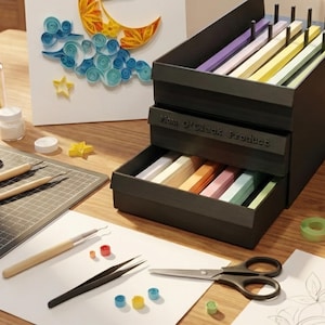 May include: A black storage unit labeled "Mom O'Clock Product" with drawers filled with colorful paper strips. Crafting tools, including scissors, tweezers, and quilling tools, are arranged on a white surface. A quilled paper art piece is in the background.