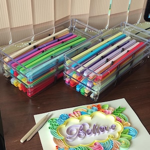 May include: Two clear plastic organizers filled with colorful paper strips. Below, a paper art piece with the word "Believe" in a decorative frame, along with a pair of craft tweezers. The scene is set on a wooden surface.