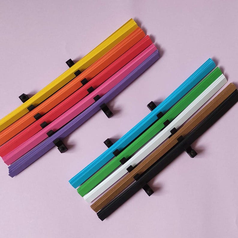10 Colors Quilling Paper Strips - High Quality 180 gsm Rainbow Palette Set DIY Kit, Typography & 3D Effect, Perfect Craft Supplies. Quilling paper strips in various widths, including 3mm, 5mm, 7mm, and 10mm. The paper is 180 GSM and comes in a pack of 100 strips. Best for - On-edge quilling, Lettering, Outline quilling and Structured designs'.The colors are black, brown, white, green, blue, purple, pink, red, orange, and yellow. The strips are arranged in a vertical row.