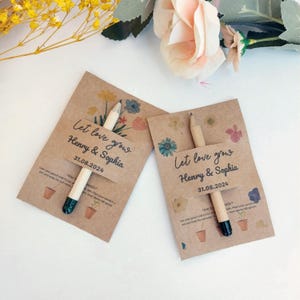 Op de afbeelding: Twee huwelijksbedankjes van kraftpapier met potloden. Elke kaart leest "Let love grow Henry & Sophia 31.08.2024" met bloemenillustraties. De potloden hebben een natuurlijke houten afwerking en een donkergroene punt.