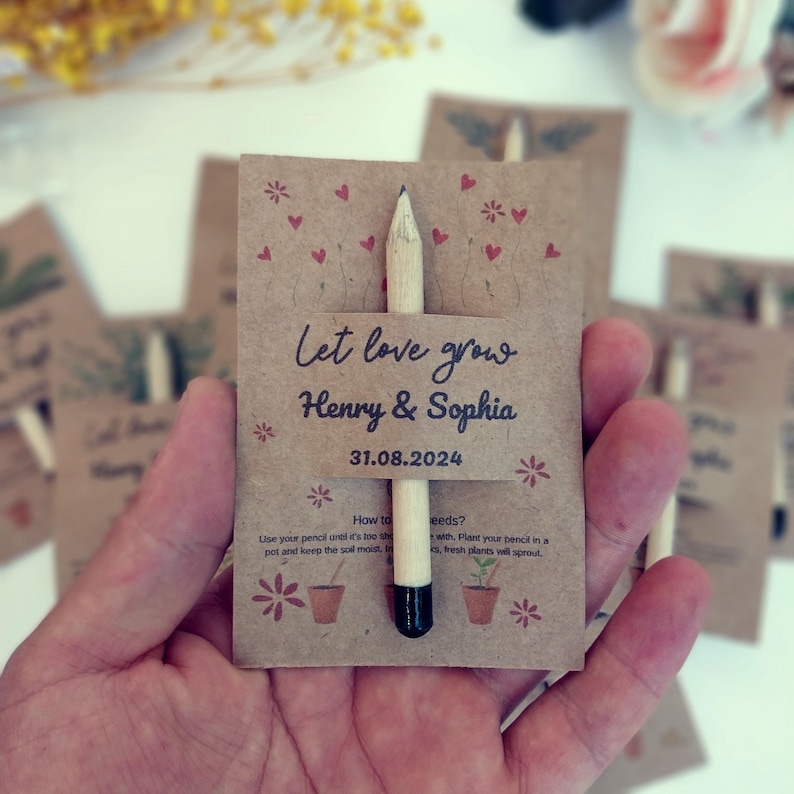 Op de afbeelding: Een trouwbedankje van kraftpapier met een potlood. De kaart leest "Let love grow Henry & Sophia 31.08.2024". De kaart is versierd met rode hartjes en bloemenillustraties. Het potlood is van natuurlijk hout met een zwarte punt.