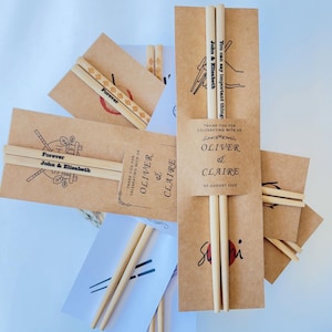 May include: A selection of wedding favours, featuring wooden chopsticks presented in brown paper sleeves. The sleeves and chopsticks are personalized with names, dates, and the word "Forever". The design is simple and elegant.