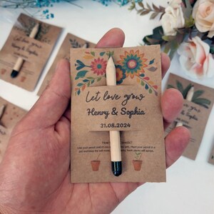 Op de afbeelding: Een trouwbedankje van kraftpapier met een potlood. De kaart leest "Let love grow Henry & Sophia 31.08.2024" met bloemmotieven. Het potlood heeft een zwarte punt en is in de kaart gestoken. Andere soortgelijke bedankjes op de achtergrond.