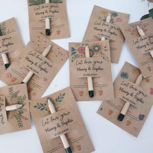 Op de afbeelding: Een verzameling kraftpapierkaarten met bloemmotieven en de tekst "Let love grow Henry & Sophia 31.08.2024." Elke kaart bevat een potlood met een groene punt. De kaarten zijn gerangschikt op een wit oppervlak.