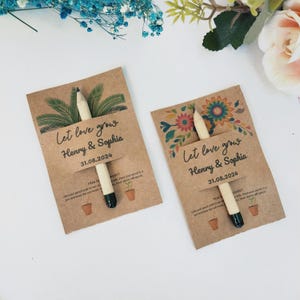 Op de afbeelding: Twee zaadpakketjes met potloden. De bruine kaarten lezen "Let love grow" met namen en een datum. E&eacute;n heeft een palmbladontwerp, de andere een bloemenontwerp. Elk heeft een potlood met een groene punt.