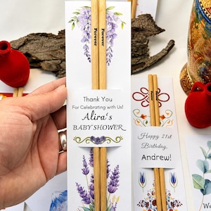 Puede incluir: Un par de palillos de bambú de color claro sujetos en una funda de papel blanco con ilustraciones de glicinias y lavanda moradas. El texto de la funda dice Thank You For Celebrating with Us! Alira's BABY SHOWER con un gráfico de rosa morada.