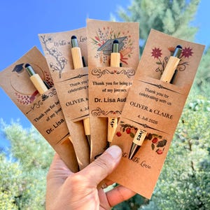 Könnte beinhalten: Vier Bambuskugelschreiber mit schwarzer Tinte und Eingabestift, befestigt an personalisierten Kraftpapierkarten. Die Karten zeigen gedruckte Abschlussmotive, florale Designs und Text mit Namen wie Dr. Lisa und Oliver & Claire.