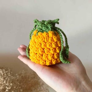 Op de afbeelding: Een handgemaakte gehaakte ananasvormige tas in helder geel met groene accenten. De tas heeft een trekkoordsluiting en wordt in een hand gehouden. Het haakwerk heeft een getextureerd, hobbelig uiterlijk.