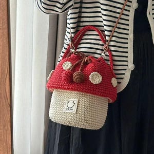 Może przedstawiać: Czerwono-beżowa szydełkowa torebka w kształcie grzyba z białymi kropkami. Torebka ma zapięcie na sznurek i małą metkę z uśmiechniętą buzią i napisem "HERE". Torebka jest noszona na brązowym pasku.