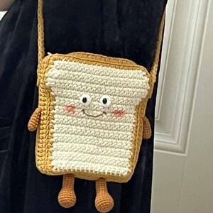 K&ouml;nnte beinhalten: Eine geh&auml;kelte braune und wei&szlig;e Tasche in Form eines Brotes mit einem l&auml;chelnden Gesicht und zwei kleinen Beinen.