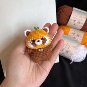 K&ouml;nnte beinhalten: Geh&auml;kelte Rotpanda-Figur in Gelb- und Braunt&ouml;nen mit wei&szlig;en Akzenten und einem kleinen gr&uuml;nen Blatt. Der Panda hat eine blaue Fliege. Die Figur wird in einer Hand gehalten. Wollkn&auml;uel sind im Hintergrund zu sehen.