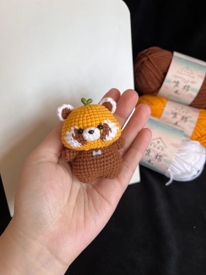 K&ouml;nnte beinhalten: Geh&auml;kelte Rotpanda-Figur in Gelb- und Braunt&ouml;nen mit wei&szlig;en Akzenten und einem kleinen gr&uuml;nen Blatt. Der Panda hat eine blaue Fliege. Die Figur wird in einer Hand gehalten. Wollkn&auml;uel sind im Hintergrund zu sehen.