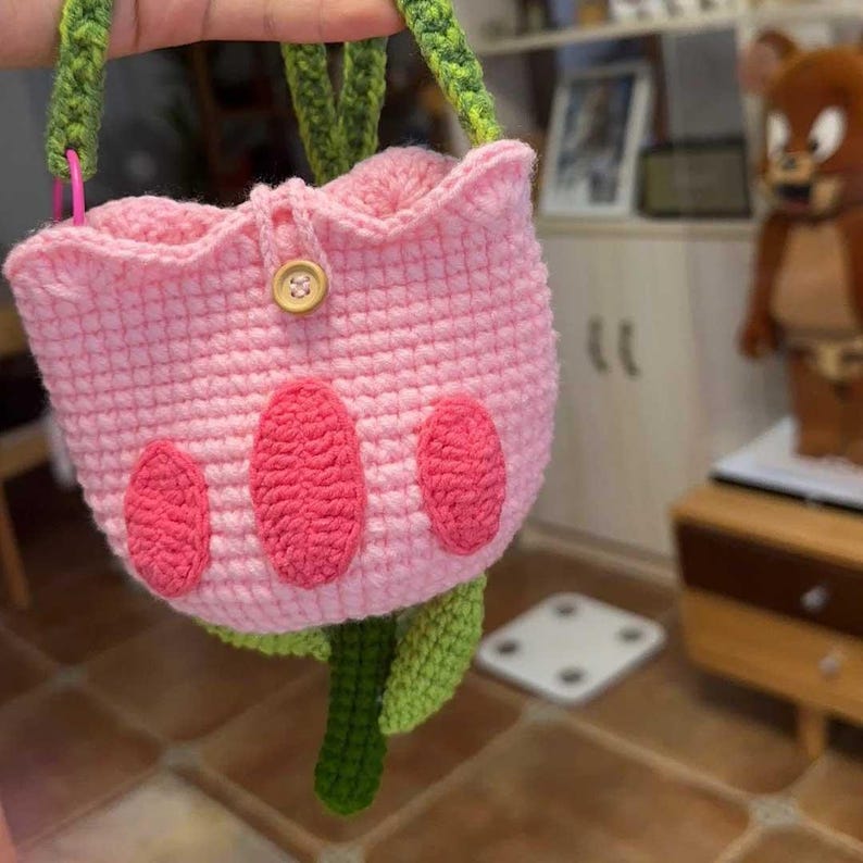 May include: A pink crocheted flower-shaped bag with green crocheted leaves and a brown button. The bag has three pink crocheted paw prints on the front.
