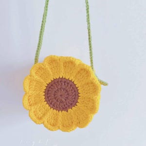 May include: A handmade, yellow crochet sunflower-shaped crossbody bag with a brown center and green strap. The bag is round with a scalloped edge, resembling a sunflower. The strap is a knitted green cord.