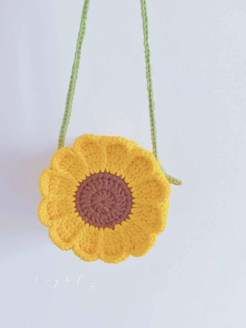 May include: A handmade, yellow crochet sunflower-shaped crossbody bag with a brown center and green strap. The bag is round with a scalloped edge, resembling a sunflower. The strap is a knitted green cord.