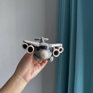 Op de afbeelding: Een handgemaakt gehaakt vliegtuigspeelgoed in grijstinten en gebroken wit. Het speelgoed heeft vleugels, een staart en vier motordetails. Het vliegtuig wordt in de hand van een persoon gehouden tegen een neutrale achtergrond.