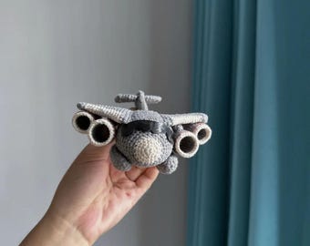 May include: A handmade crochet airplane toy in shades of gray and off-white. The toy features wings, a tail, and four engine details. The airplane is held in a person's hand against a neutral background.
