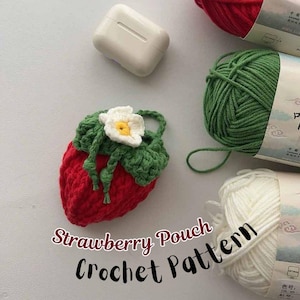 Może przedstawiać: Ręcznie robiona czerwona szydełkowa torebka w kształcie truskawki z zielonymi liśćmi i białym kwiatem. Torebka znajduje się obok białego etui na AirPody i kłębków wełny. Tekst "Strawberry Pouch Crochet Pattern" jest również na zdjęciu.