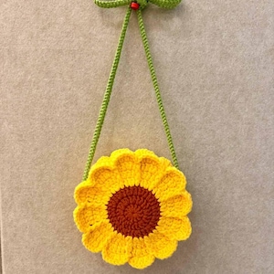 May include: A handmade crochet sunflower bag with a brown center and yellow petals. The bag has a green strap and a green bow at the top. The bag is approximately 7 inches in diameter.