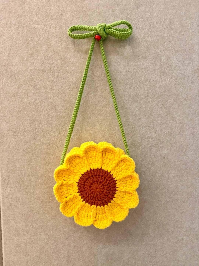 May include: A handmade crochet sunflower bag with a brown center and yellow petals. The bag has a green strap and a green bow at the top. The bag is approximately 7 inches in diameter.