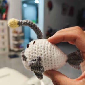 K&ouml;nnte beinhalten: Ein handgefertigtes H&auml;kelfisch-Spielzeug. Das Spielzeug ist wei&szlig; mit grauen Flossen und einem grau-gelben K&ouml;der. Der Anglerfisch hat kleine braune Augen.