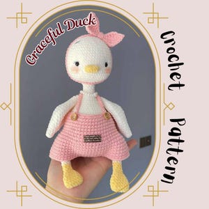 May include: A crocheted duck toy with a pink dress and a pink bow. The duck is white with yellow feet and a yellow beak. The toy is being held in a hand. The text "Graceful Duck Crochet Pattern" is written on the image.
