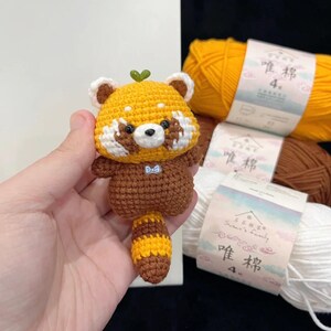 K&ouml;nnte beinhalten: Handgefertigte geh&auml;kelte Rotpanda-Figur mit gelbem Kopf, braunen Akzenten und gestreiftem Schwanz. Das kleine Pl&uuml;schtier hat ein gr&uuml;nes Blatt auf dem Kopf und eine blaue Fliege. Die Figur wird in einer Hand gehalten.