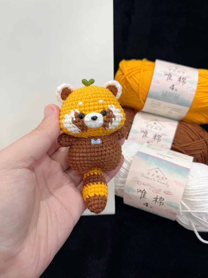 K&ouml;nnte beinhalten: Handgefertigte geh&auml;kelte Rotpanda-Figur mit gelbem Kopf, braunen Akzenten und gestreiftem Schwanz. Das kleine Pl&uuml;schtier hat ein gr&uuml;nes Blatt auf dem Kopf und eine blaue Fliege. Die Figur wird in einer Hand gehalten.