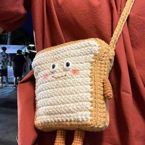 K&ouml;nnte beinhalten: Eine geh&auml;kelte Tasche in Brotform mit einem l&auml;chelnden Gesicht und zwei kleinen Beinen. Die Tasche ist aus braunem und wei&szlig;em Garn gefertigt und hat einen braunen Riemen.