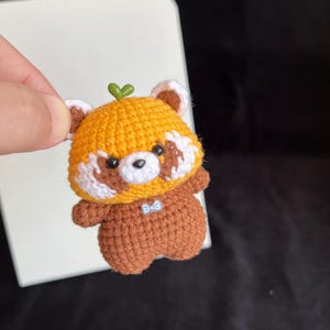 K&ouml;nnte beinhalten: Handgefertigte geh&auml;kelte Rotpanda-Figur. Der Rotpanda hat einen gelben Kopf mit wei&szlig;en und braunen Akzenten, einen braunen K&ouml;rper und ein kleines gr&uuml;nes Blatt oben. Er hat eine kleine blaue Fliege.