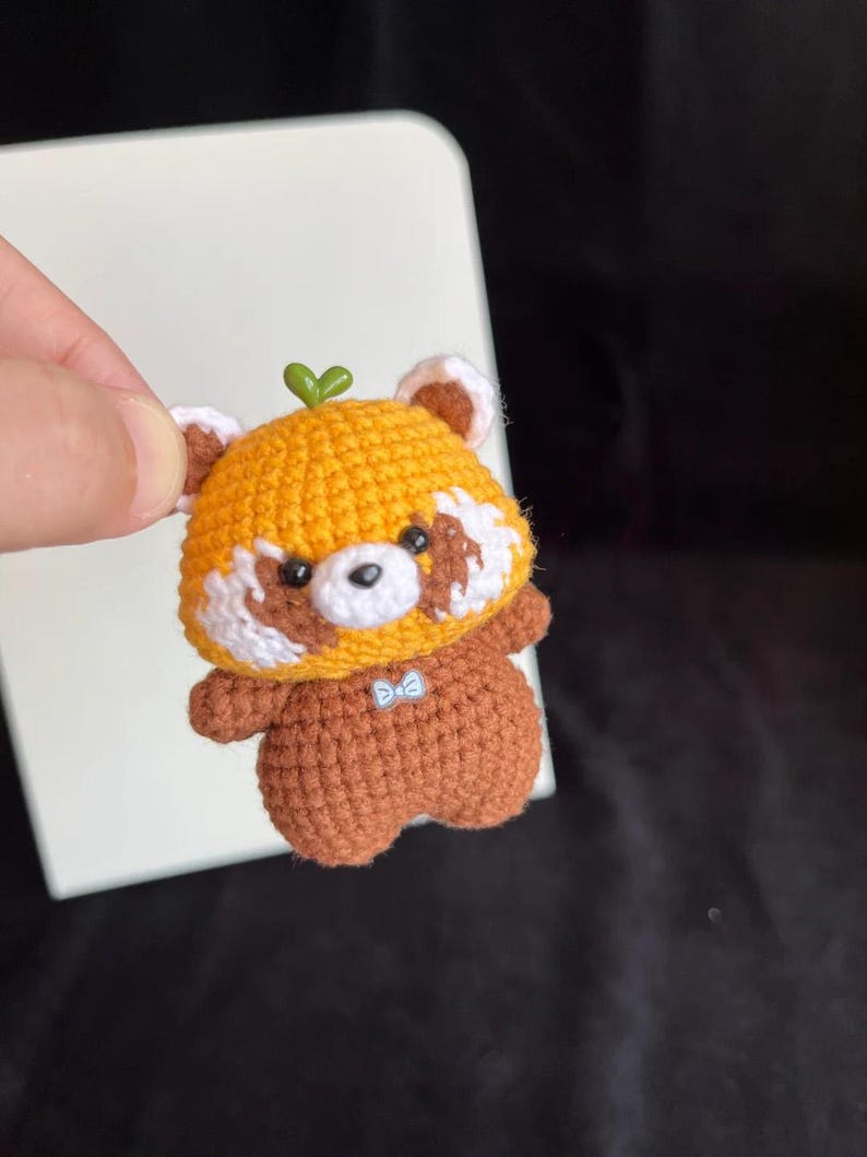 K&ouml;nnte beinhalten: Handgefertigte geh&auml;kelte Rotpanda-Figur. Der Rotpanda hat einen gelben Kopf mit wei&szlig;en und braunen Akzenten, einen braunen K&ouml;rper und ein kleines gr&uuml;nes Blatt oben. Er hat eine kleine blaue Fliege.