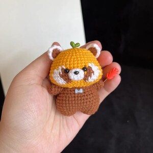 K&ouml;nnte beinhalten: Handgefertigte geh&auml;kelte Rotpanda-Figur. Der Panda hat einen gelben Kopf mit wei&szlig;en Akzenten, einen braunen K&ouml;rper und ein kleines gr&uuml;nes Blatt oben. Er h&auml;lt einen kleinen orangefarbenen Gegenstand. Die Figur ist ca. 7.6 cm gro&szlig;.