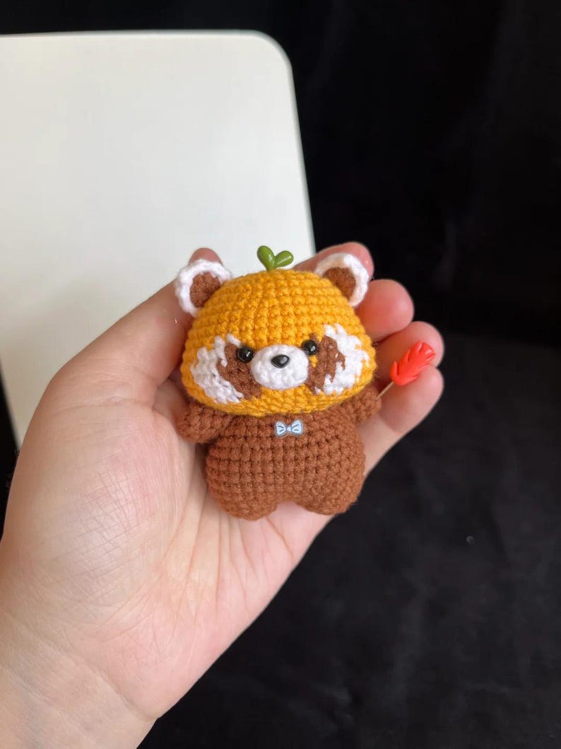 K&ouml;nnte beinhalten: Handgefertigte geh&auml;kelte Rotpanda-Figur. Der Panda hat einen gelben Kopf mit wei&szlig;en Akzenten, einen braunen K&ouml;rper und ein kleines gr&uuml;nes Blatt oben. Er h&auml;lt einen kleinen orangefarbenen Gegenstand. Die Figur ist ca. 7.6 cm gro&szlig;.
