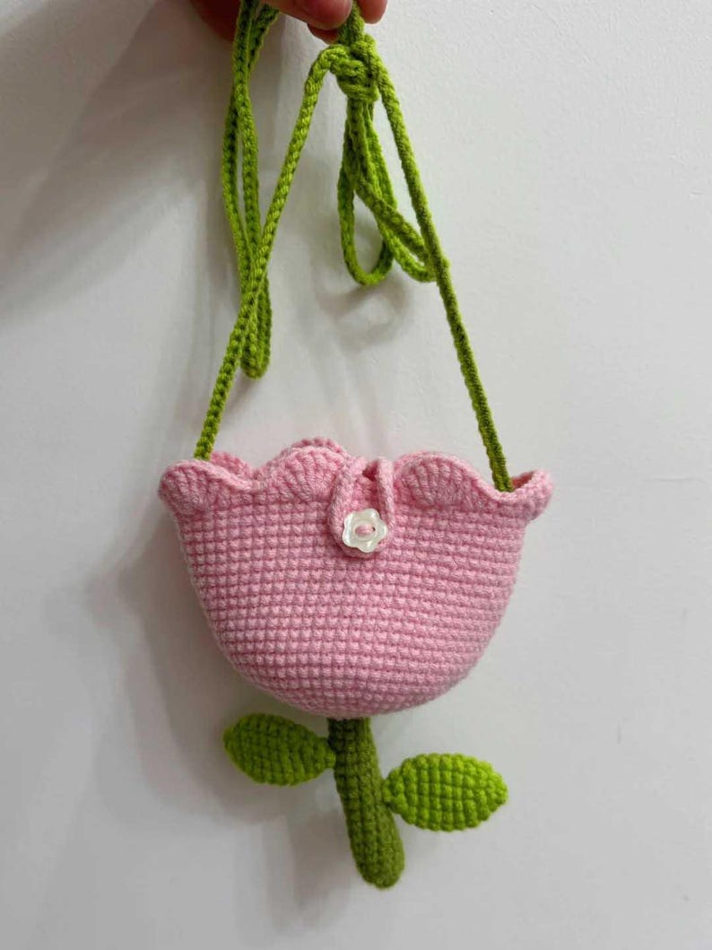 May include: A pink crocheted tulip-shaped bag with a green crocheted stem and leaves. The bag has a small white button on the front.