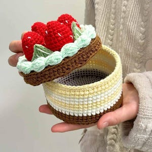 May include: A crocheted round container with a lid shaped like a strawberry cake. The lid features red strawberries, green leaves, and a light green frosting border. The container has brown, yellow, and white stripes.