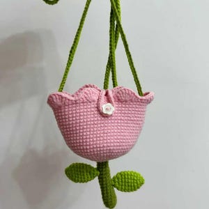 May include: A pink crocheted flower-shaped bag with a green strap and leaves. The bag has a white button on the front.
