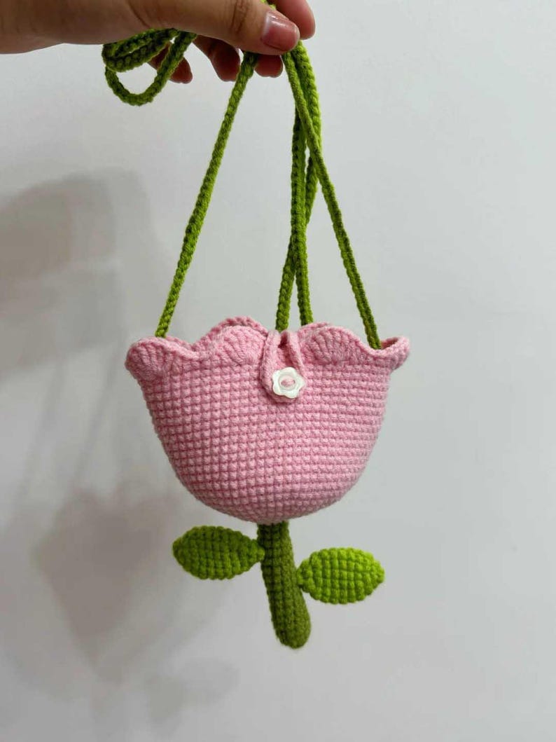 May include: A pink crocheted flower-shaped bag with a green strap and leaves. The bag has a white button on the front.